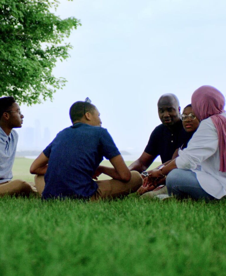 Ashley, a Black hijabi, sits on the grass in a park with her husband, two teen sons, and her teen daughter