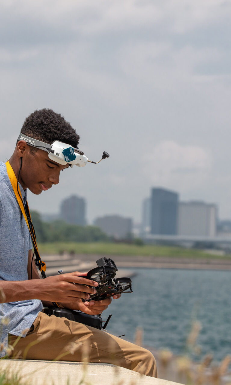 Yusef, a young adult, looking at a drone with his equipment on his head. The background is an amphitheater leading to the water with a cityscape in the far background.