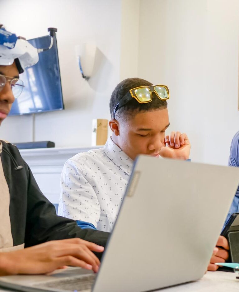 Two teen Black boys and their sister work at laptops and tablets. Their sister wears a hijab