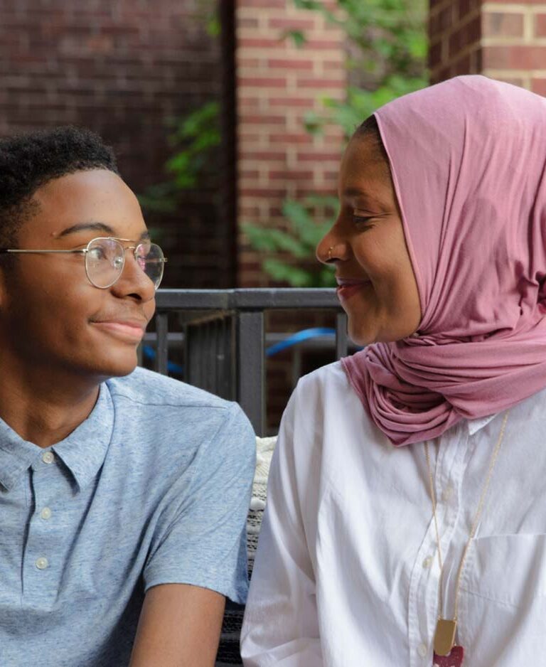 A mother in a hijab looks at her teenage son in glasses, and they're both smiling.