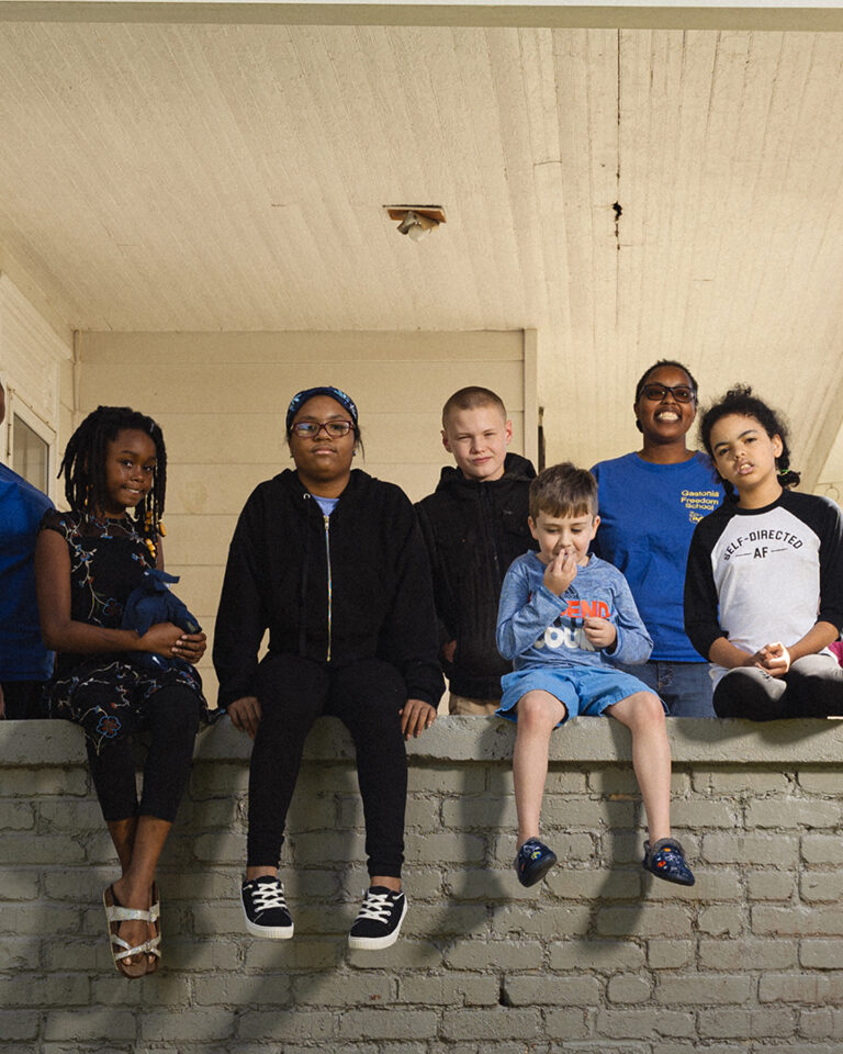 8 children smile together, sitting on a porch railing. They're flanked by two Black women