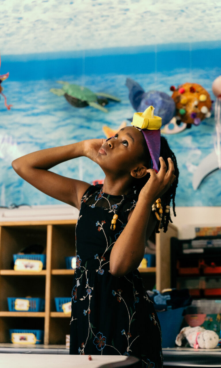 A girl putting on a headband and looking up at the sky while in a classroom.