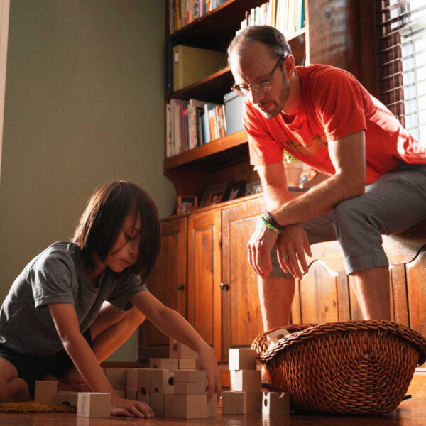 A kid playing with big wooden blocks while his father looks on.