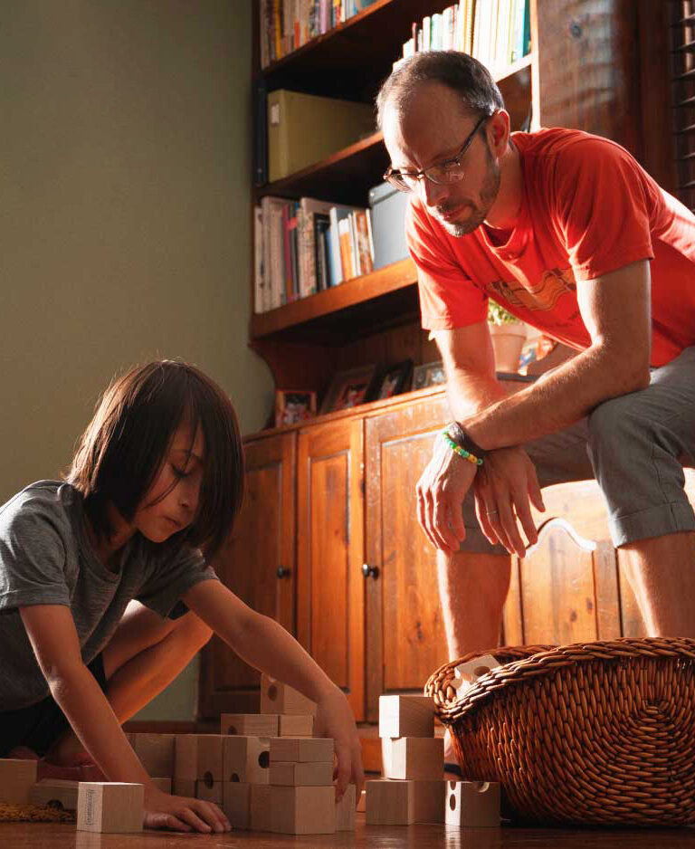 A kid playing with big wooden blocks while his father looks on.